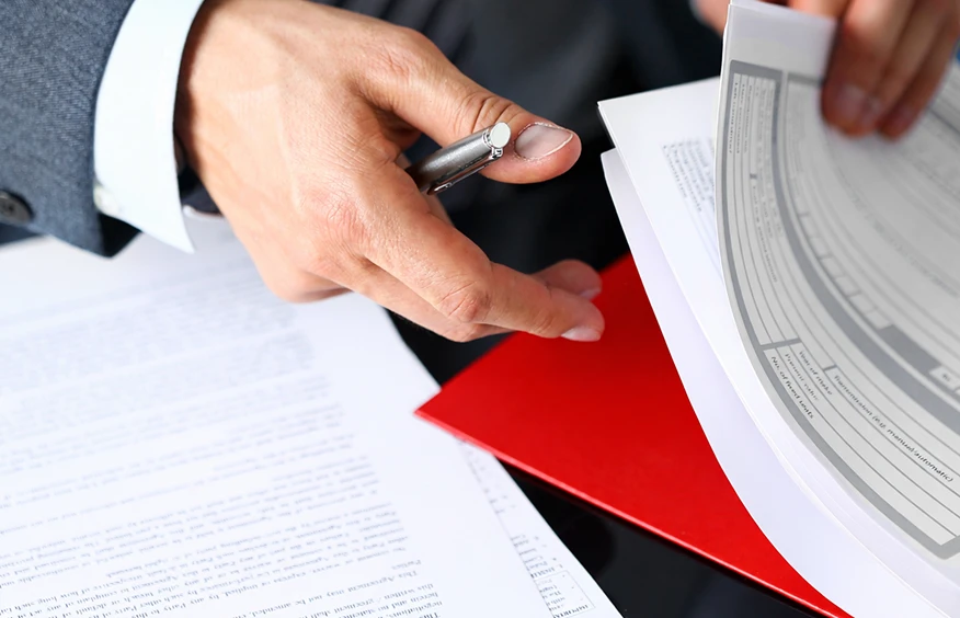 Человек в деловом костюме держит ручку и документы, внимательно изучая бумаги.